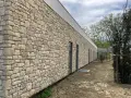 Un mur extérieur en pierres naturelles irrégulières, avec des fenêtres modernes et un toit blanc. Le sol est en terre battue.