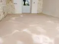 Sol fraîchement ragréé, l'épaisseur du ragréage intérieur est visible sur le sol brut. Les murs en pierre et la porte blanche annoncent une rénovation.