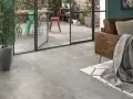 Un salon moderne avec un canapé en cuir marron et un plaid gris. Le sol est recouvert d'un carrelage usé que faire ? Une grande baie vitrée donne sur un patio avec des chaises colorées.