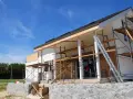 Une maison en construction, avec des échafaudages et une échelle, en cours d'agrandissement. Le ciel bleu est parsemé de nuages légers.
