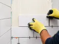 Pose-t-on coller du carrelage avec du ciment ? Oui, un carreleur pose du carrelage blanc sur un mur avec des croisillons.