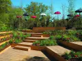 Un jardin en pente moderne avec des terrasses en bois, des parterres de fleurs rouges et des parasols colorés.