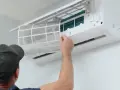 Un homme retire un filtre à air sale d'une unité de climatisation murale, étape essentielle de l'entretien climatisation maison pour un air sain.
