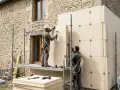 Deux ouvriers installent des panneaux isolants sur un mur de pierre. Ils utilisent des échafaudages et des outils pour fixer le matériau, assurant ainsi le meilleur isolant thermique pour la bâtisse.