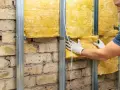 Un ouvrier installe une laine de verre jaune sur une structure métallique fixée à un mur de briques, améliorant l'isolation phonique.