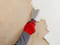 Main tenant utilise un grattoir pour décoller papier peint ancien sur un mur.