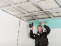 Un ouvrier réalise le schema montage placo plafond, fixant une plaque verte sur une structure métallique.