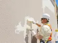 Un ouvrier en casque blanc applique un nouvel enduit extérieur sur un enduit existant, à l'aide de deux truelles.