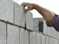 Main d'un maçon plaçant une parpaing pour monter un mur en parpaing de 2m. Le fil à plomb guide la pose.