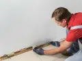 Un homme en gants gris est en train d'enlever la plinthe de carrelage le long d'un mur blanc.