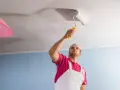 Un homme en salopette blanche et t-shirt rose utilise un rouleau pour peindre un plafond. Il est concentré sur sa tâche, montrant comment peindre un plafond.