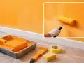 Un rouleau de peinture jaune crée des traces de rouleau à la lumière sur un mur orange. Matériel de peinture à proximité.