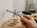Main d'un bricoleur tenant un cheville métallique pour charge lourde sur carreau de plâtre. Divers chevilles et outils en arrière-plan.