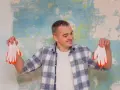 Homme souriant tenant des gants blancs à bouts orange devant un mur texturé. Il pense au prix peinture m2 au black pour son projet.