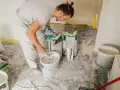 Un ouvrier prépare de l'enduit béton cellulaire dans un seau avec un mélangeur. Des sacs de poudre sont à proximité.