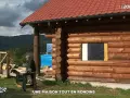 Une maison en rondins de bois en construction, avec un homme travaillant à proximité. Le toit est rouge et le paysage est montagneux.