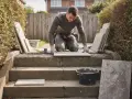 Un ouvrier pose des dalles sur un escalier extérieur en béton. Des outils et des matériaux sont à proximité.