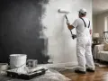 Un peintre en salopette blanche est en train de repeindre un mur de couleur sombre en blanc, utilisant un rouleau.