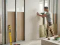 Un homme utilise une perceuse pour monter une cloison en placo sur une structure métallique. Des matériaux d'isolation sont visibles.
