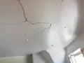 Un plafond qui s'effrite, révélant des fissures sombres et des morceaux de plâtre tombés. La lumière douce éclaire les imperfections.