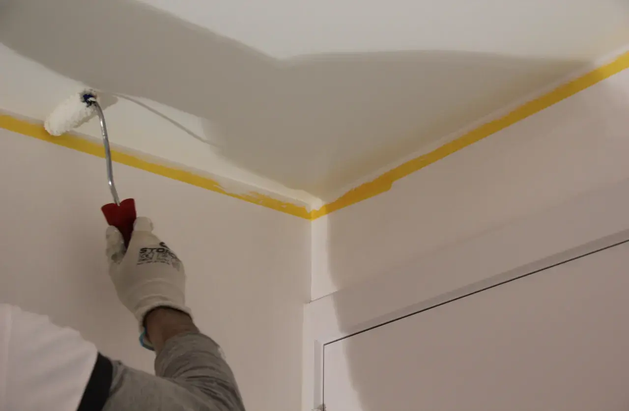 Maintenant, vous savez comment peindre un plafond : une main gantée utilise un rouleau pour peindre le coin d'un plafond, protégé par du ruban de masquage jaune.