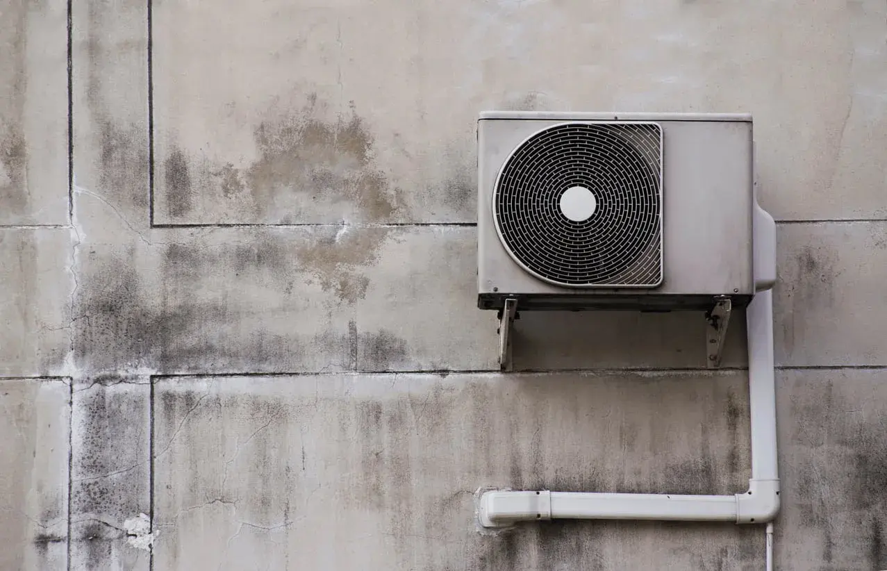 Unité extérieure d'une pompe à chaleur air air fixée sur un mur en béton fissuré et taché.