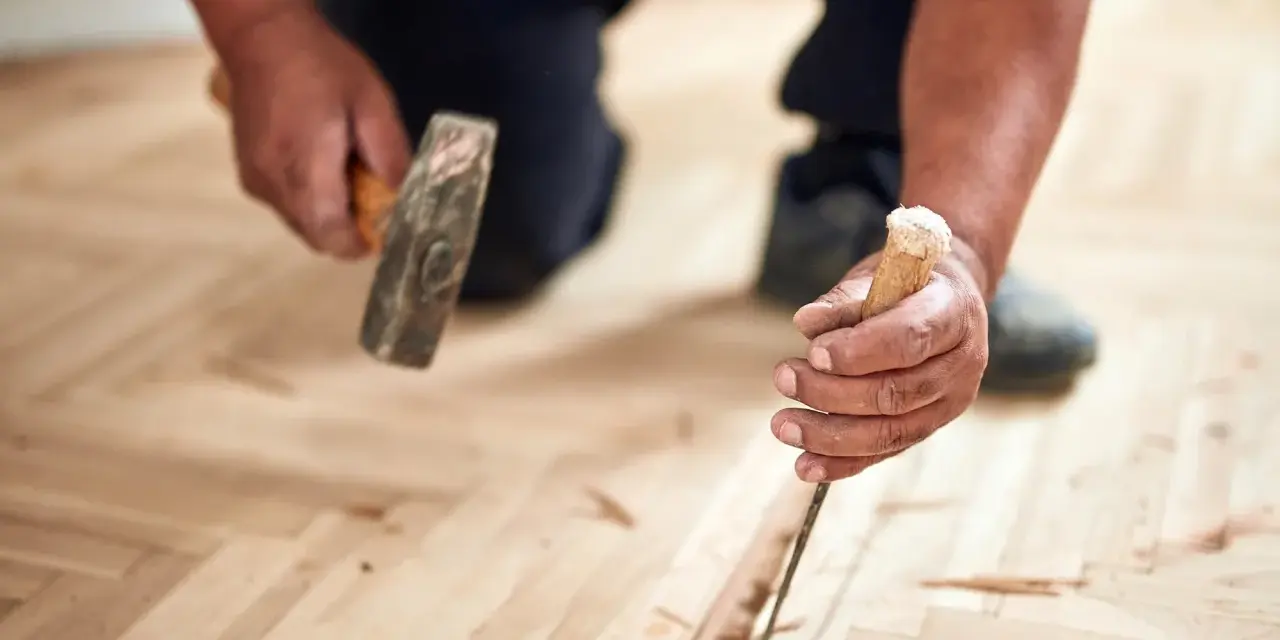 Un artisan utilise un ciseau et un marteau pour changer une lame de parquet flottant endommagée.