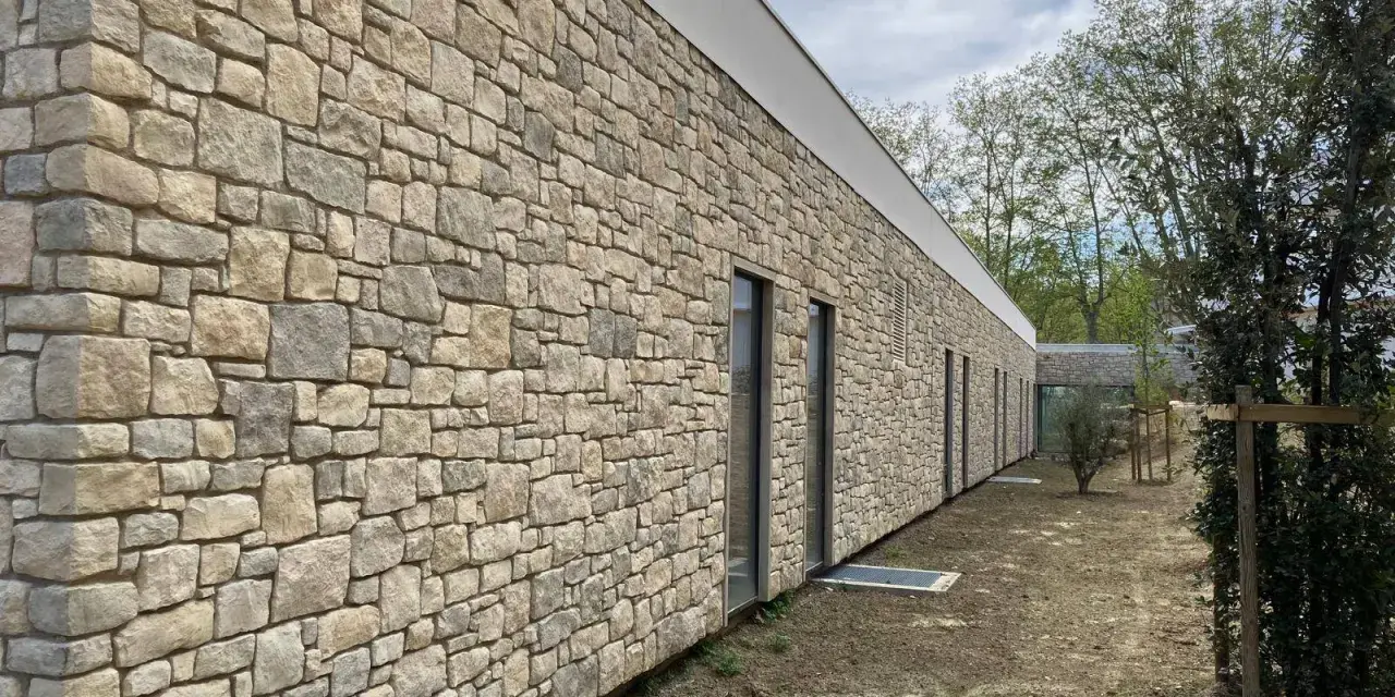 Un mur extérieur en pierres naturelles irrégulières, avec des fenêtres modernes et un toit blanc. Le sol est en terre battue.
