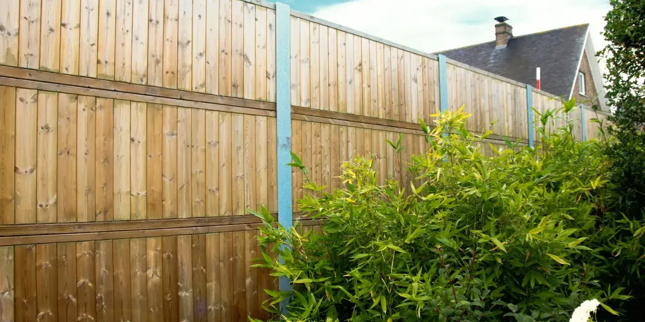Clôture anti bruit en bois, haute et solide, protégeant un jardin verdoyant. Le ciel bleu et les nuages ajoutent une touche de sérénité.