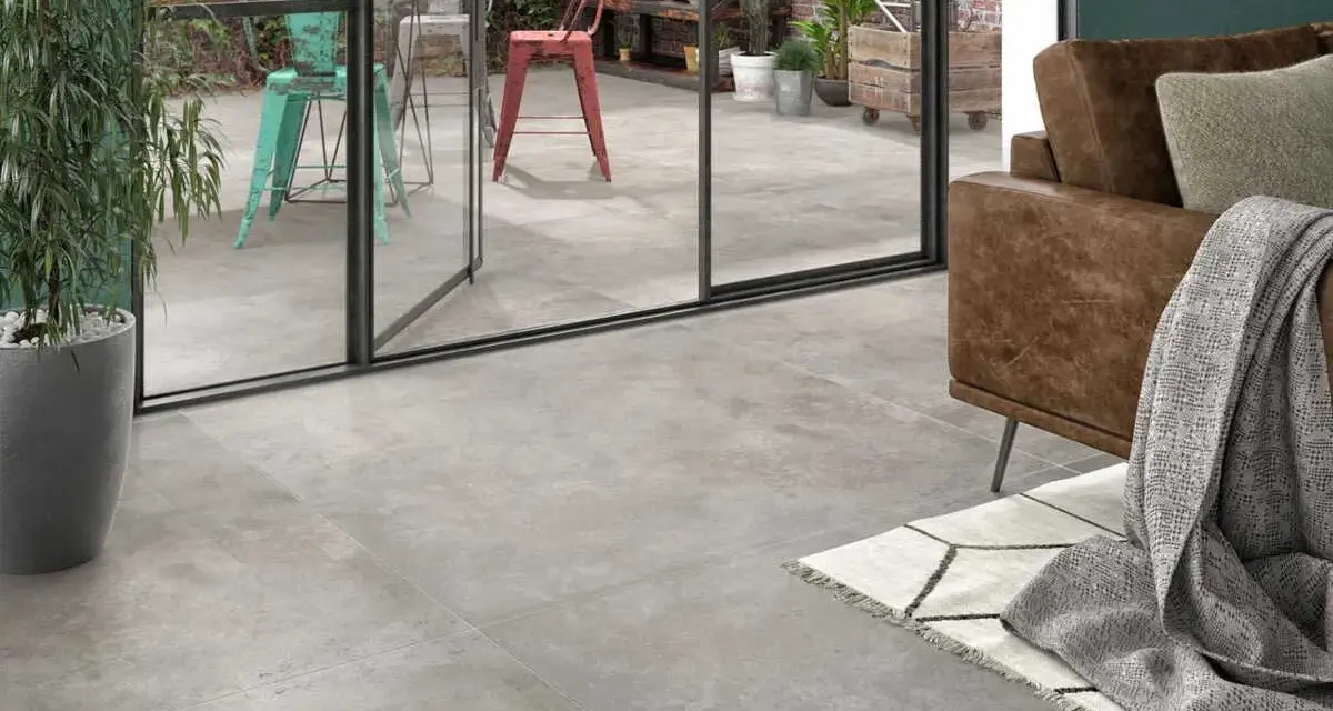 Un salon moderne avec un canapé en cuir marron et un plaid gris. Le sol est recouvert d'un carrelage usé que faire ? Une grande baie vitrée donne sur un patio avec des chaises colorées.