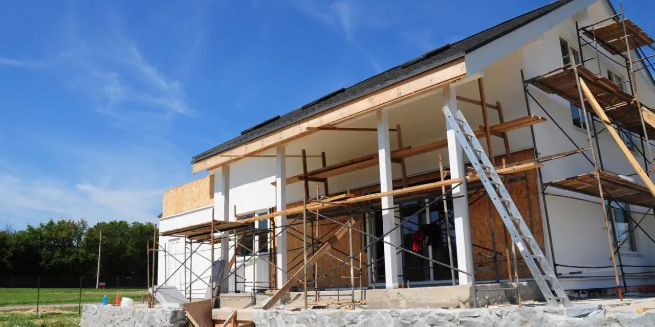 Une maison en construction, avec des échafaudages et une échelle, en cours d'agrandissement. Le ciel bleu est parsemé de nuages légers.