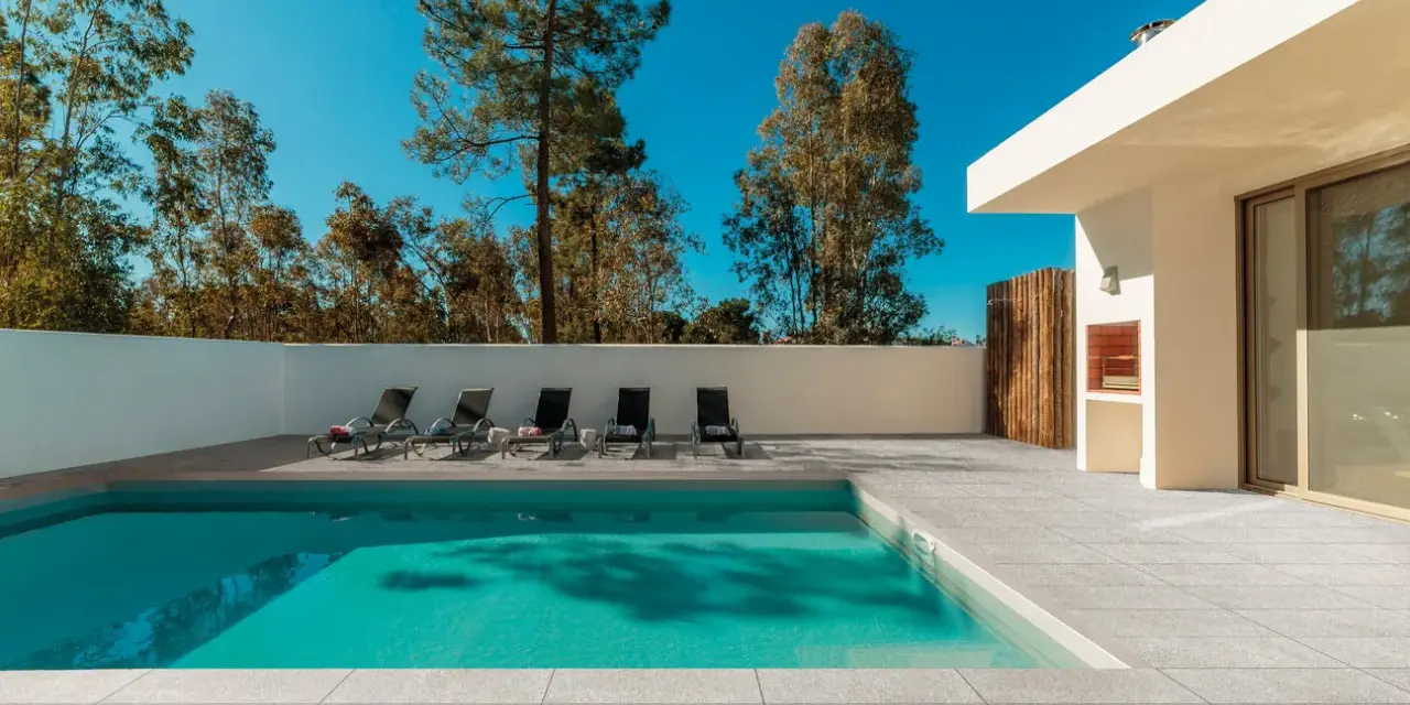Une piscine moderne avec un sol en carrelage gris clair, des chaises longues et un barbecue extérieur sous un ciel bleu éclatant.