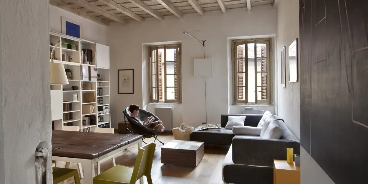 Salon avec poutres apparentes au plafond, une personne lisant dans un fauteuil, canapé, table et étagères.