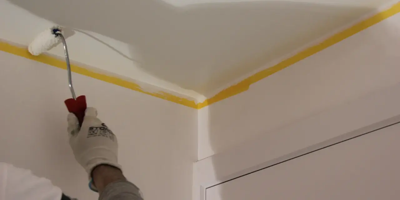 Maintenant, vous savez comment peindre un plafond : une main gantée utilise un rouleau pour peindre le coin d'un plafond, protégé par du ruban de masquage jaune.