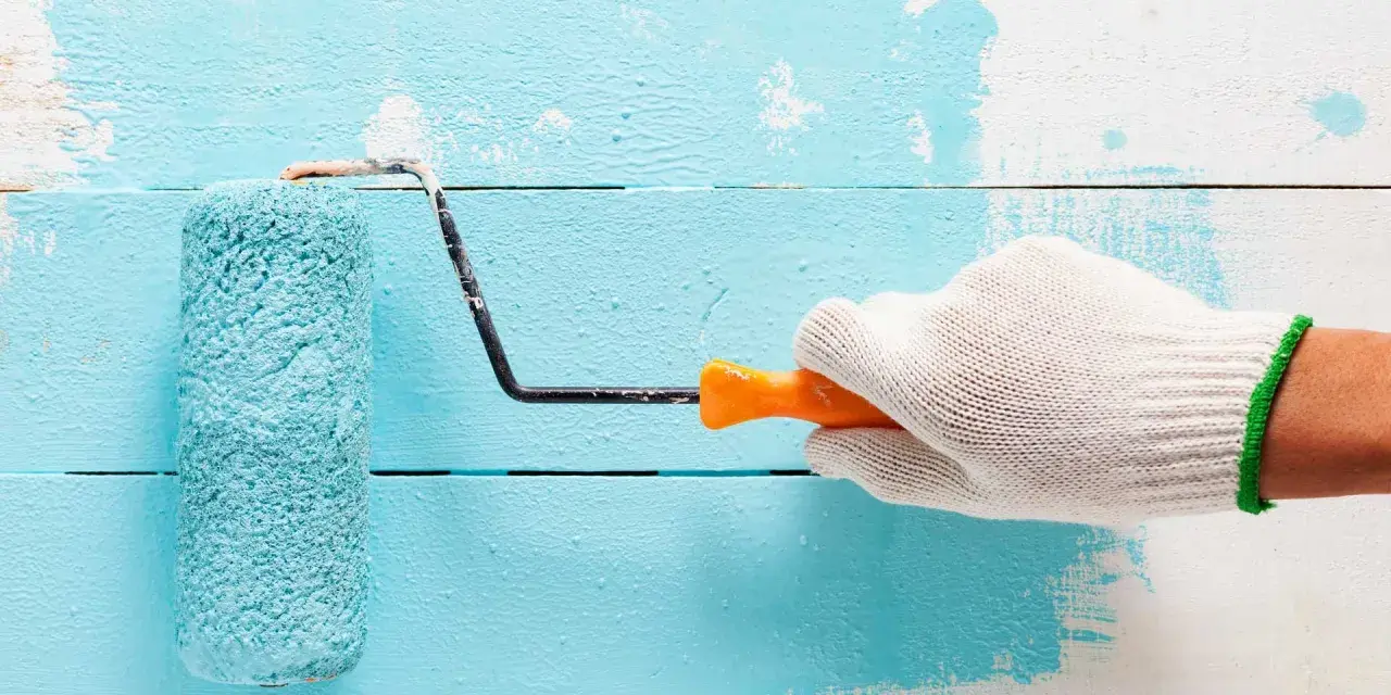 Main tenant une main gantée, un rouleau applique une peinture bleu turquoise sur un mur en bois blanc.