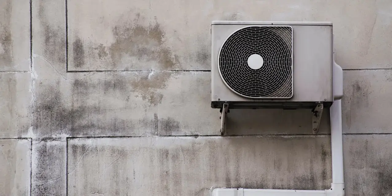 Unité extérieure d'une pompe à chaleur air air fixée sur un mur en béton fissuré et taché.