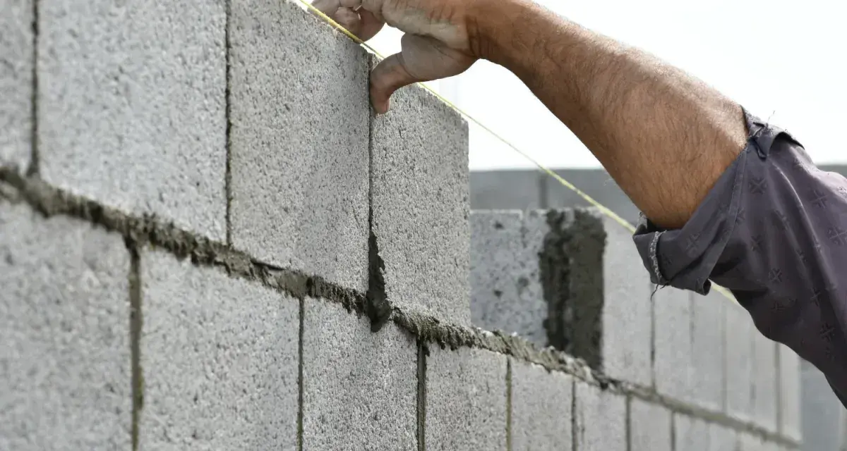 Main d'un maçon plaçant une parpaing pour monter un mur en parpaing de 2m. Le fil à plomb guide la pose.