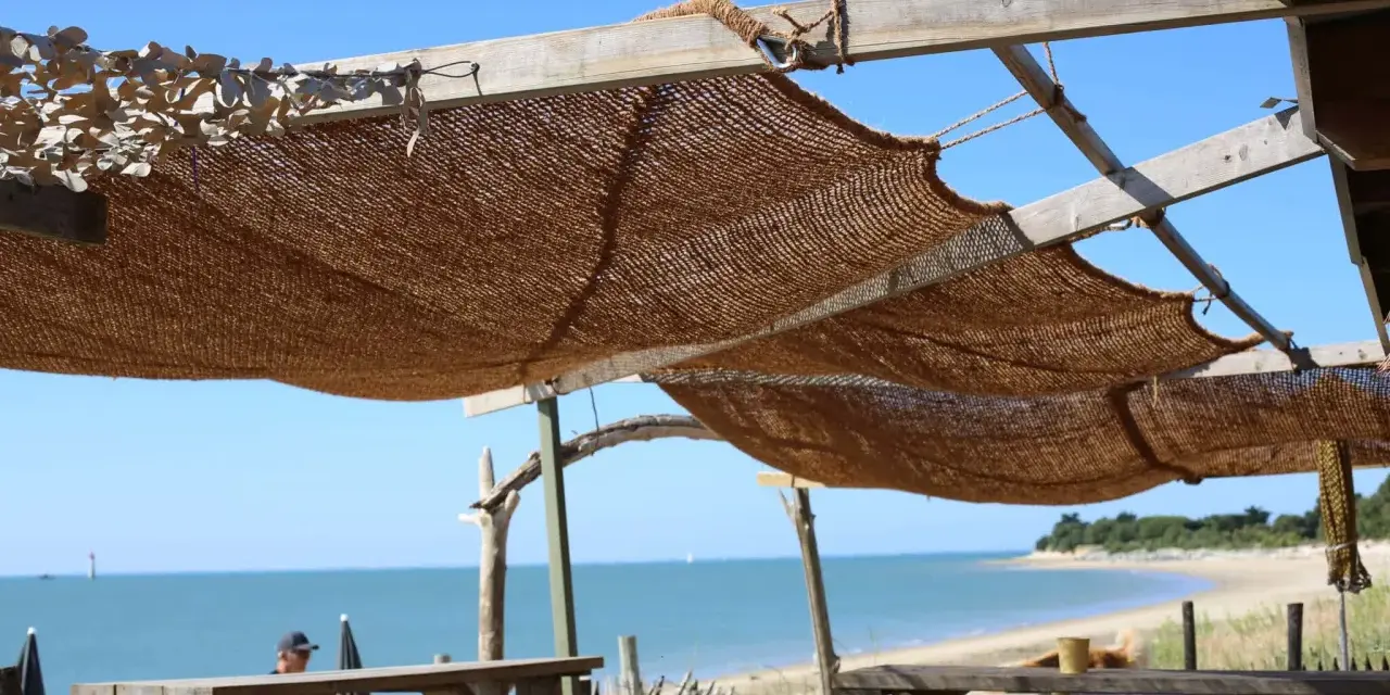 Installation d'une voile d'ombrage en toile de jute sur une structure en bois, offrant une vue sur la mer et la plage.