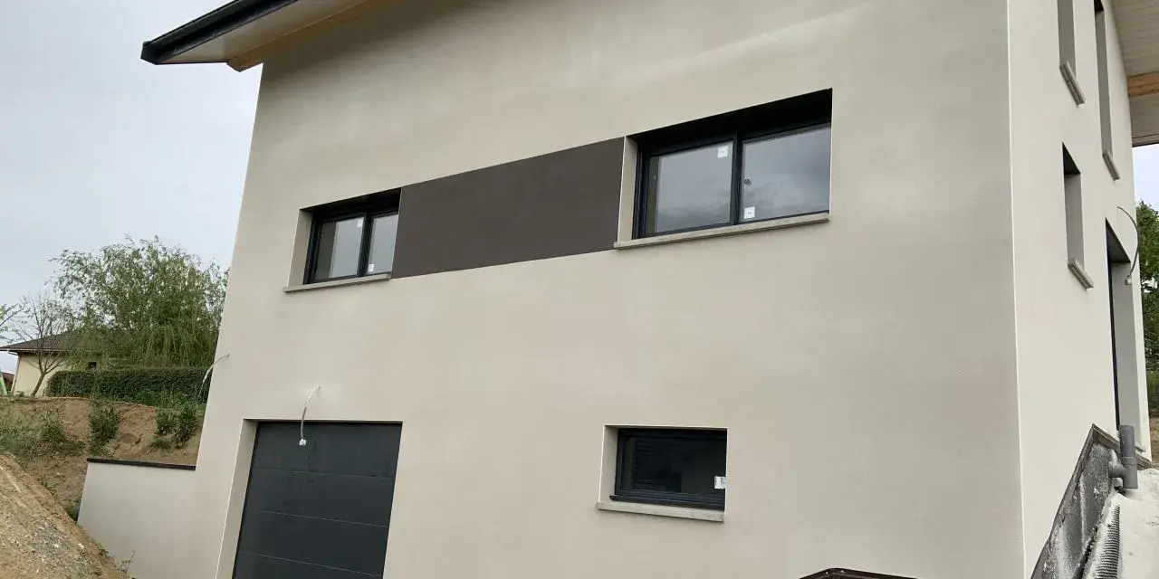Maison moderne avec enduit neuf, une trace blanche discrète sur le crépi clair. Fenêtres noires et garage gris.