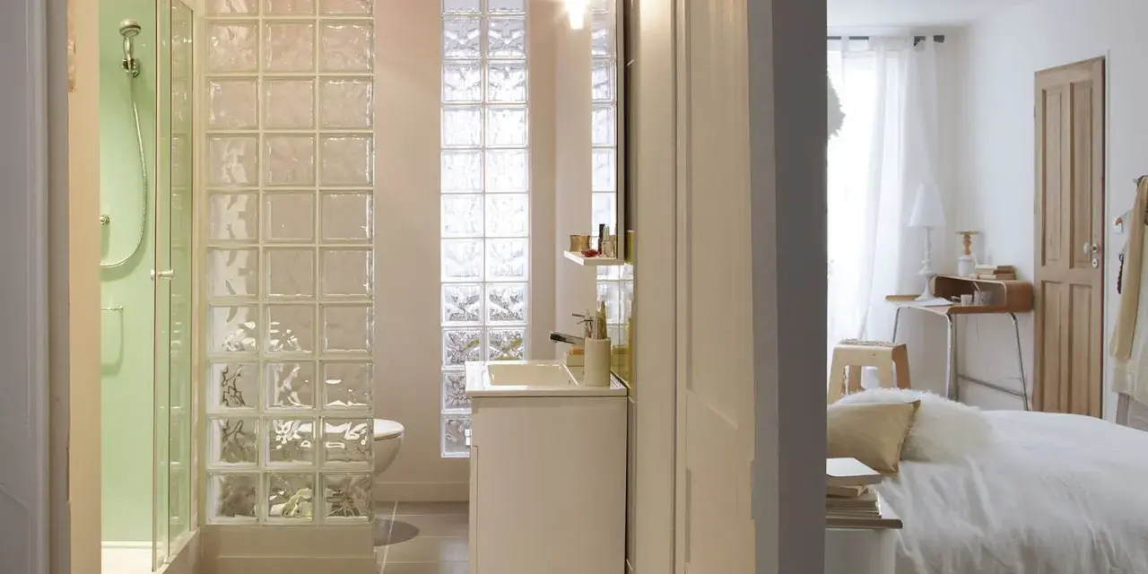 Salle de bain moderne avec douche, lavabo et toilettes, séparée de la chambre par un mur en brique de verre.