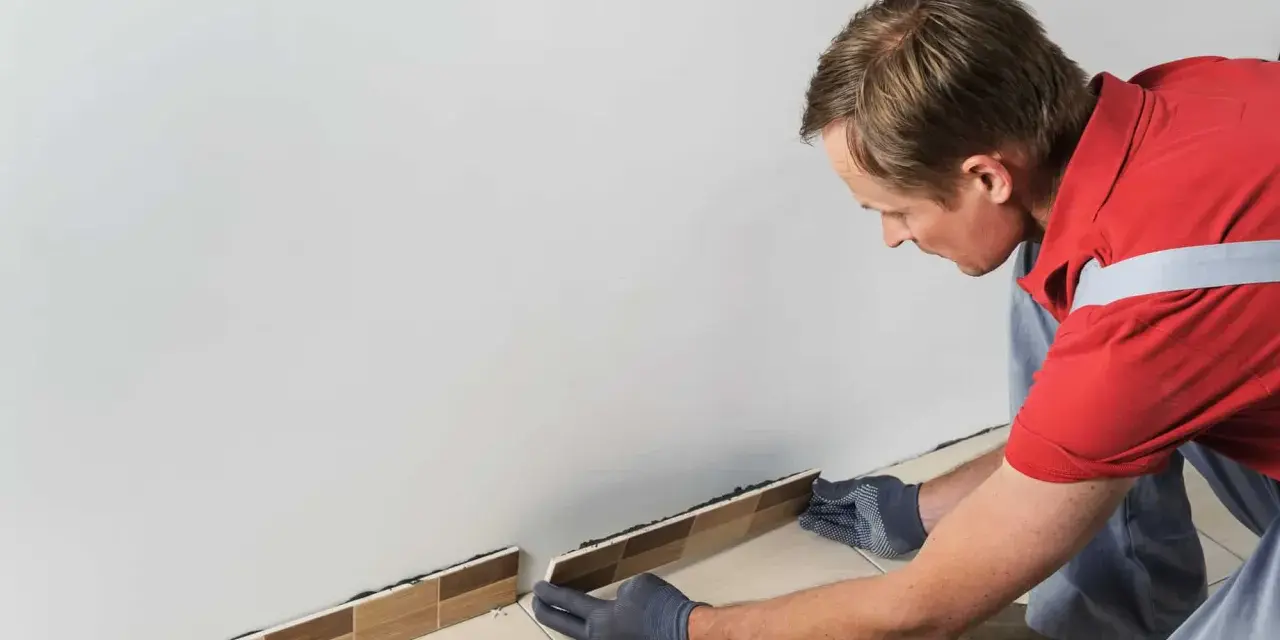 Un homme en gants gris est en train d'enlever la plinthe de carrelage le long d'un mur blanc.