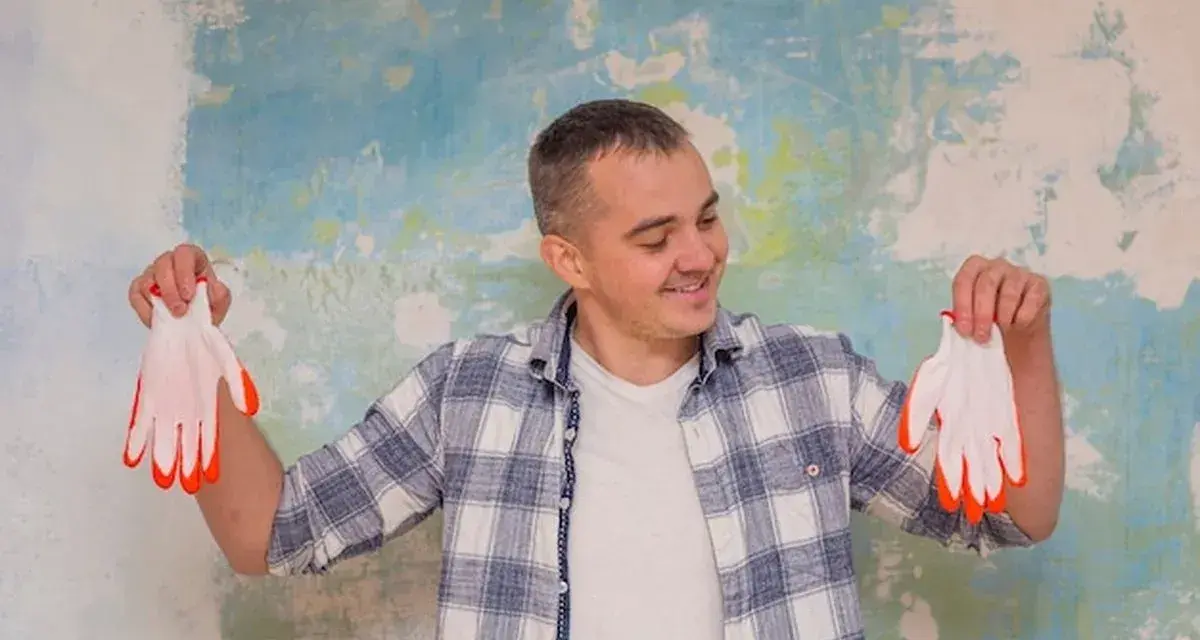 Homme souriant tenant des gants blancs à bouts orange devant un mur texturé. Il pense au prix peinture m2 au black pour son projet.