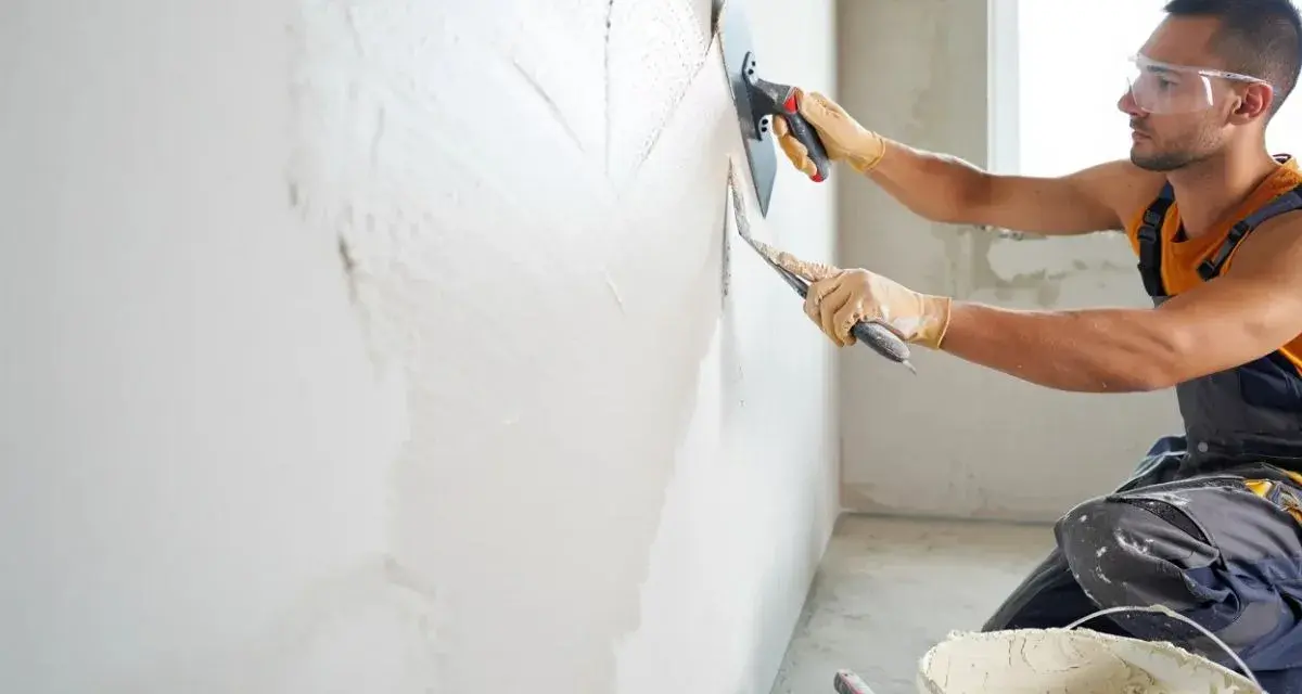 Un ouvrier applique un enduit sur un mur humide avec une truelle et une taloche. Le travail progresse, laissant une surface lisse.
