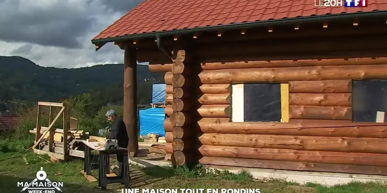 Une maison en rondins de bois en construction, avec un homme travaillant à proximité. Le toit est rouge et le paysage est montagneux.