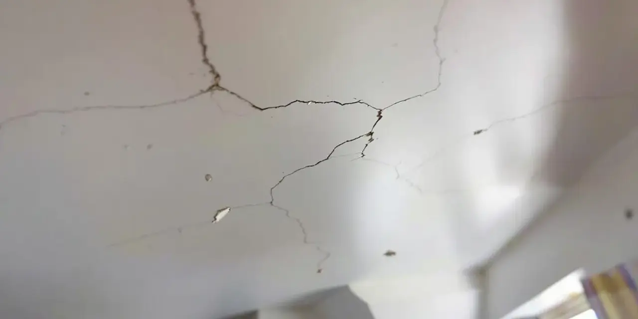 Un plafond qui s'effrite, révélant des fissures sombres et des morceaux de plâtre tombés. La lumière douce éclaire les imperfections.