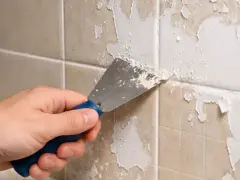 Main tenant enlever peinture sur carrelage avec une spatule. La peinture s'écaille, révélant le carrelage beige en dessous.