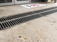 Un panneau au sol indique "Sip iPhone 115€". Un caniveau en métal longe un sol en béton. Le béton drainant prix m2 est une solution pour évacuer l'eau.