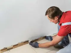Un homme en gants gris est en train d'enlever la plinthe de carrelage le long d'un mur blanc.