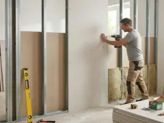 Un homme utilise une perceuse pour monter une cloison en placo sur une structure métallique. Des matériaux d'isolation sont visibles.