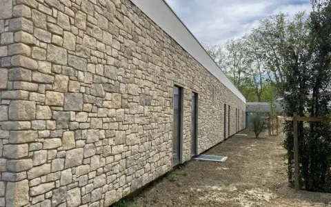 Un mur extérieur en pierres naturelles irrégulières, avec des fenêtres modernes et un toit blanc. Le sol est en terre battue.